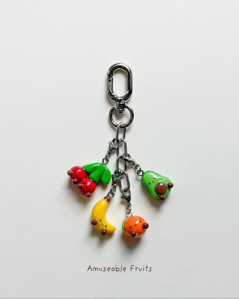 May include: A silver keychain with a lobster clasp holding charms shaped like a cherry, avocado, banana, and orange. The charms are colorful and have smiling faces. The text "Amuseable Fruits" is at the bottom.