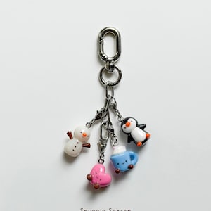 May include: A silver keychain with four winter-themed charms: a snowman, a pink heart, a blue mug with whipped cream, and a penguin. The charms are attached to a silver clasp. The text "Snuggle Season" is at the bottom.