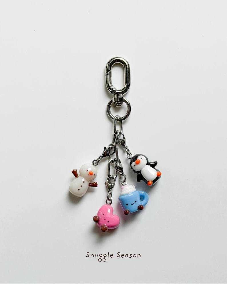 May include: A silver keychain with four winter-themed charms: a snowman, a pink heart, a blue mug with whipped cream, and a penguin. The charms are attached to a silver clasp. The text "Snuggle Season" is at the bottom.
