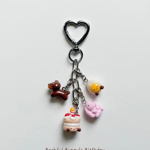 May include: A keychain with a heart-shaped clasp and several charms. Charms include a brown dachshund, a cake with a lit candle, a fried egg, and a pink bunny. The text "Bashful Bunny's Birthday" is at the bottom.