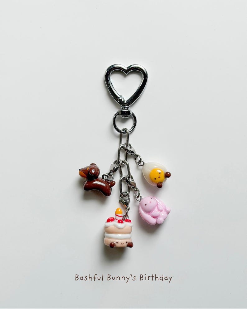 May include: A keychain with a heart-shaped clasp and several charms. Charms include a brown dachshund, a cake with a lit candle, a fried egg, and a pink bunny. The text "Bashful Bunny's Birthday" is at the bottom.