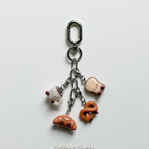 May include: A silver keychain with five miniature food charms: a coffee cup, a slice of toast, a pretzel, a croissant, and a bread roll. The charms are attached to a chain and a clasp. The text "Coffee & Crumbs" is at the bottom.