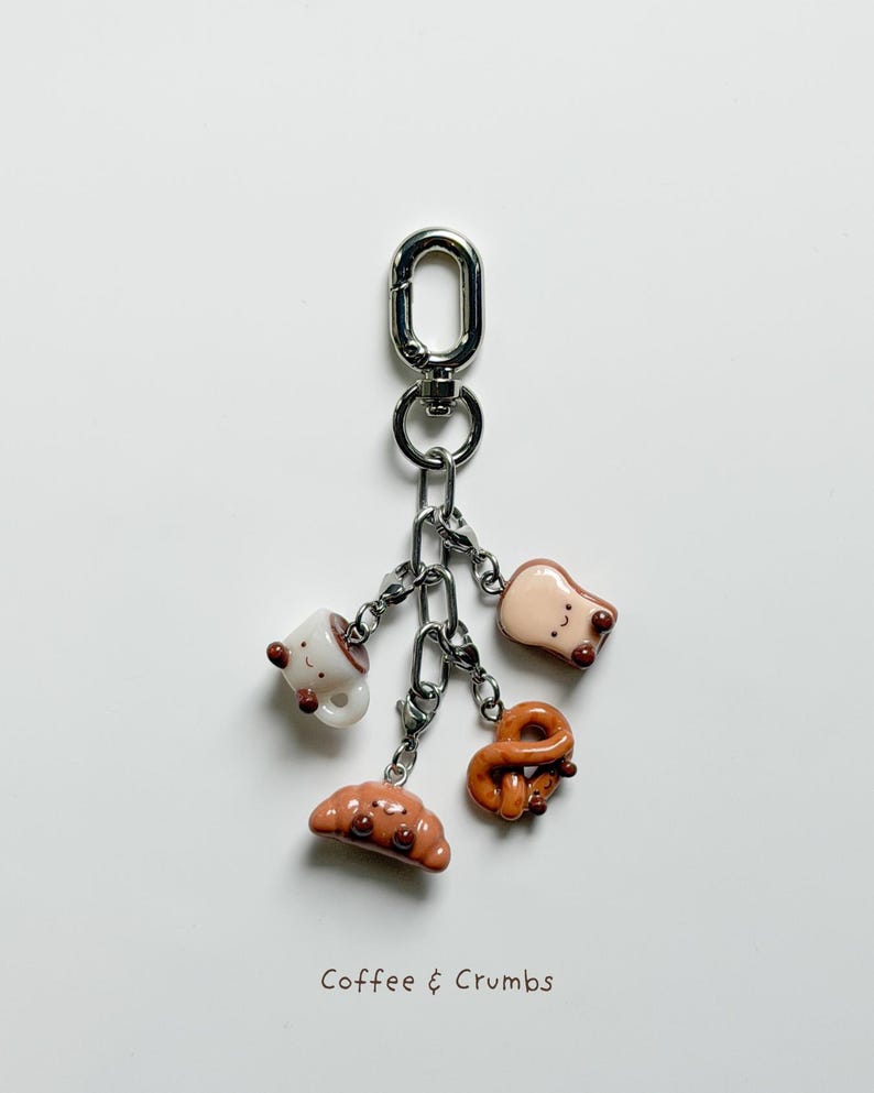 May include: A silver keychain with five miniature food charms: a coffee cup, a slice of toast, a pretzel, a croissant, and a bread roll. The charms are attached to a chain and a clasp. The text "Coffee & Crumbs" is at the bottom.