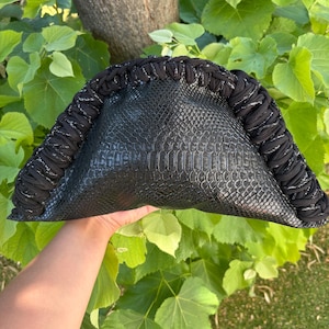 Peut inclure: Une pochette semi-circulaire présentant une texture en faux cuir de serpent noir sur le corps principal. Le bord incurvé est encadré par une épaisse bordure en tissu noir tressé intégrant de subtils fils métalliques argentés.