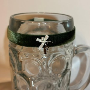 May include: A clear glass beer stein with a decorative green velvet band around the top. A small silver charm with a bow and the letter "F" hangs from the band. The stein has a handle and a textured pattern.