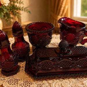 May include: A set of deep red pressed glass tableware featuring a butter dish with a lid, a creamer pitcher, a sugar bowl, and two shakers. Each piece displays a textured diamond and circle pattern with scalloped edges and a glossy finish.