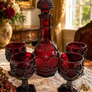May include: A deep red glass decanter with a matching stopper and four matching stemmed glasses. The set features a textured hobnail and geometric pattern across the surface of the glass, with a hexagonal base on the decanter and the glasses.
