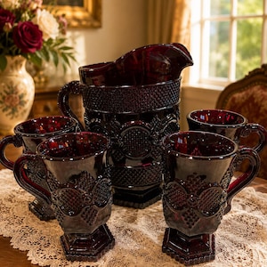 May include: A deep red glass pitcher and four matching mugs featuring a textured diamond pattern and circular motifs. The set includes a large pouring vessel with a curved handle and four pedestal-based mugs, all crafted from dark ruby-colored glass.