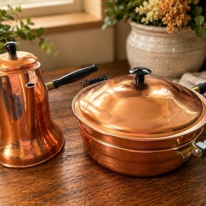 May include: A polished copper kettle with a black handle and a matching copper pot with a lid and black side handles. Both items feature a shiny metallic finish and dark, ribbed handles, showcasing a classic design for kitchen use.