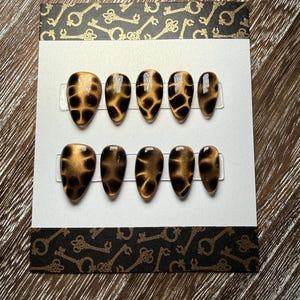 May include: A set of ten almond-shaped press-on nails. The nails have a brown and black tortoiseshell pattern. The nails are displayed on a white card with decorative black and gold key-patterned borders.
