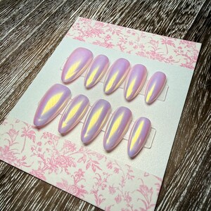 May include: A set of iridescent, oval-shaped false nails in shades of pink and yellow. The nails are arranged on a pink floral patterned card. The nails have a glossy finish and are ready for application.
