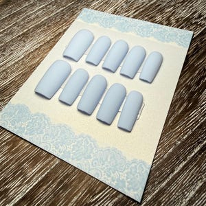 May include: A set of ten light blue matte finish press-on nails arranged on a decorative card with a light blue lace border. The nails are oval-shaped and evenly spaced, showcasing a solid, pastel color. The background is a wooden surface.
