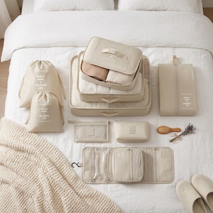 Peut inclure: Ensemble d'organisateurs et d'accessoires de voyage beiges, comprenant des cubes d'emballage, des sacs à cordon, un sac à chaussures et un organiseur de toilette. Le texte "WISH U HAVE A COLORFUL LIFE" est imprimé sur certains articles. Une brosse à cheveux en bois et une brindille de lavande sont également présentes.