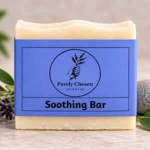 May include: A rectangular off-white soap bar wrapped in a blue paper band. The band features a circular logo with a hand holding a sprig, the text Purely Chosen SKINCARE, and the words Soothing Bar printed in a bold black sans-serif font.