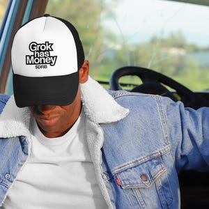 May include: A black and white trucker hat with the text "Grok has Money" in a bold, stylized font. The hat is worn by a person in a denim jacket with a white sherpa collar, inside a vehicle.