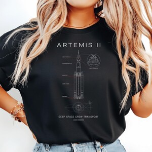 Op de afbeelding: Zwart T-shirt met een gedetailleerd diagram van de Artemis II-missie, inclusief de raket en de onderdelen. De tekst "ARTEMIS II" staat bovenaan, met "DEEP SPACE CREW TRANSPORT" onderaan. Thema ruimteverkenning.