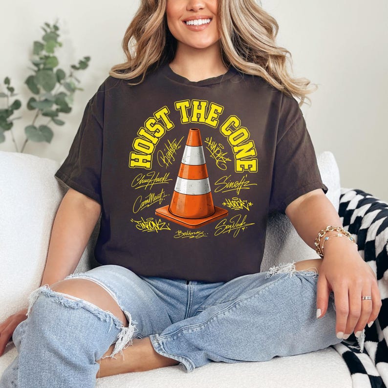 Op de afbeelding: Bruin t-shirt met de gele tekst "HOIST THE CONE" en een oranje en witte verkeerskegelafbeelding. Het ontwerp bevat verschillende handtekeningen rond de kegel.