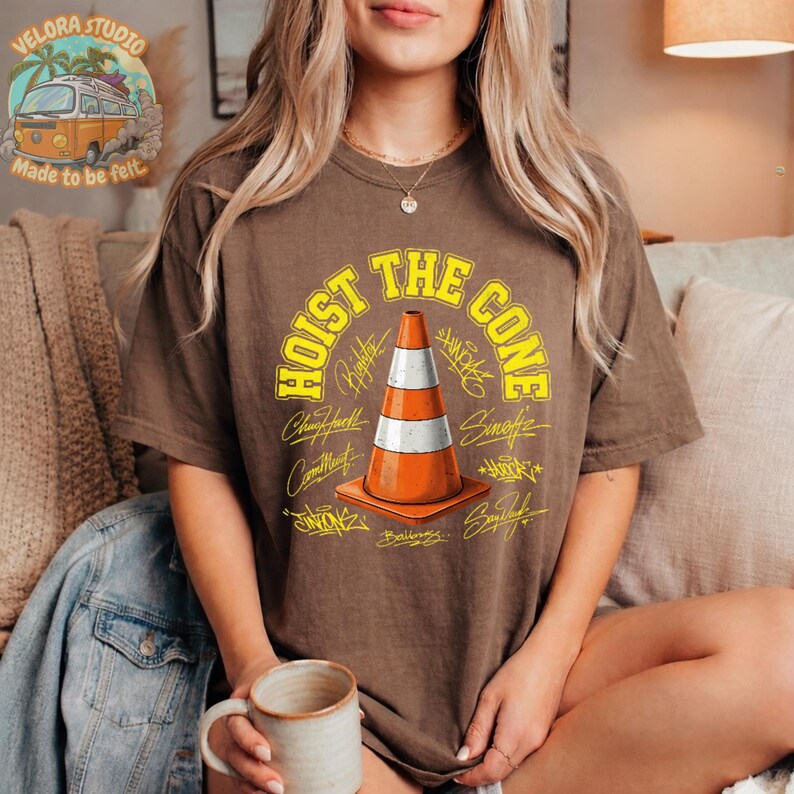 Op de afbeelding: Bruin t-shirt met de gele tekst "HOIST THE CONE" en een oranje en witte verkeerskegelafbeelding. Het shirt heeft een relaxte pasvorm en is gemaakt van een zacht materiaal. Het ontwerp bevat verschillende handtekeningen rond de kegel.