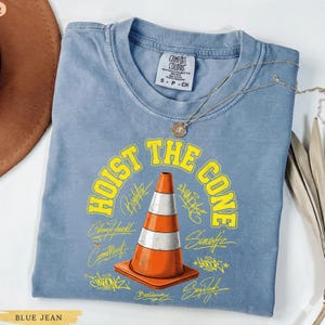 Op de afbeelding: Blauw Comfort Colors T-shirt met een afbeelding van een oranje en witte verkeerskegel en de gele woorden "HOIST THE CONE". Het shirt heeft ook verschillende handtekeningen en het Comfort Colors label.