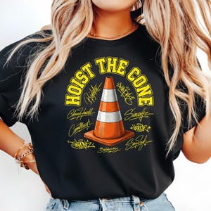 Op de afbeelding: Zwart t-shirt met een gele en oranje verkeerskegelafbeelding met de tekst "HOIST THE CONE" in het geel. De kegel heeft witte en oranje strepen en is omringd door handtekeningen.