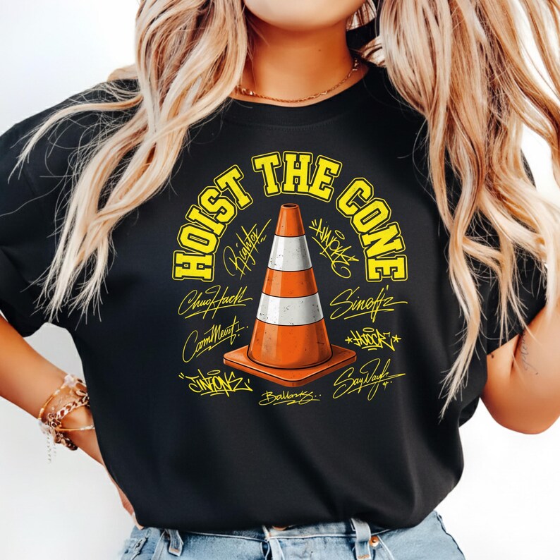 Op de afbeelding: Zwart t-shirt met een gele en oranje verkeerskegelafbeelding met de tekst "HOIST THE CONE" in het geel. De kegel heeft witte en oranje strepen en is omringd door handtekeningen.