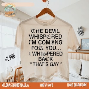 May include: Cream-colored t-shirt with the text "THE DEVIL WHISPERED I'M COMING FOR YOU... I WHISPERED BACK 'THAT'S GAY'" in black. The shirt is hanging on a wooden hanger. The image includes a logo and text at the bottom.