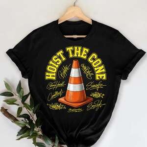 Op de afbeelding: Zwart T-shirt met een afbeelding van een oranje en witte verkeerskegel. De tekst "HOIST THE CONE" is geel. Het ontwerp bevat handtekeningen rond de kegel.