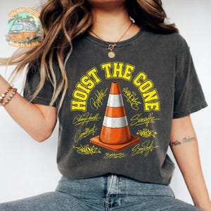Op de afbeelding: Donkergrijs t-shirt met de gele tekst "HOIST THE CONE" en een oranje en witte verkeerskegelafbeelding. Het shirt bevat ook handtekeningen en het Velora Studio-logo.