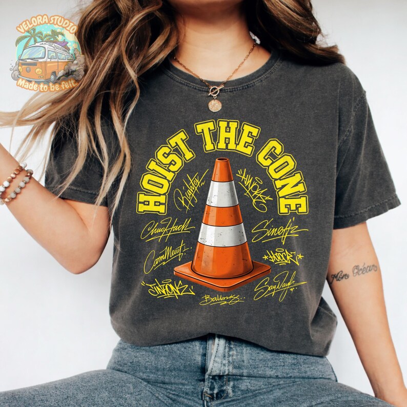 Op de afbeelding: Donkergrijs t-shirt met de gele tekst "HOIST THE CONE" en een oranje en witte verkeerskegelafbeelding. Het shirt bevat ook handtekeningen en het Velora Studio-logo.