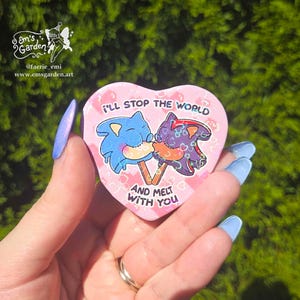 May include: A heart-shaped pin with a pink background featuring two character-shaped popsicles, one blue and one dark purple, with the text I'LL STOP THE WORLD AND MELT WITH YOU printed in black. The surface has a holographic heart pattern.