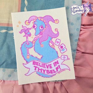May include: A rectangular fabric patch featuring a blue fuzzy worm character wearing a purple jester hat with bells. The character holds a matching jester scepter and is surrounded by small playing cards. A banner below reads BELIEVE IN THYSELF!