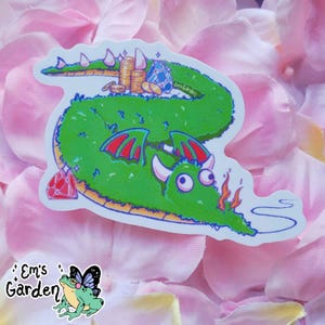 May include: A quirky sticker of a green dragon with red wings, white horns, and large eyes. The dragon is decorated with coins, gems, and a ruby. The sticker includes the text "Em's Garden" and a frog with a butterfly design.