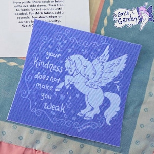 May include: A square fabric patch in shades of blue featuring a winged horse illustration and the text 'your kindness does not make you weak'. The design includes decorative swirls and stars, with a small 'Em's Garden' logo in the corner.
