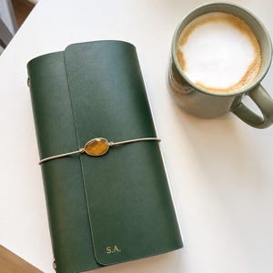 May include: A dark green leather journal with a gold-coloured clasp and the initials "S.A." embossed on the cover. A cup of coffee with a foamy top sits nearby on a white surface. The journal is closed and secured with a thin, silver-coloured band.