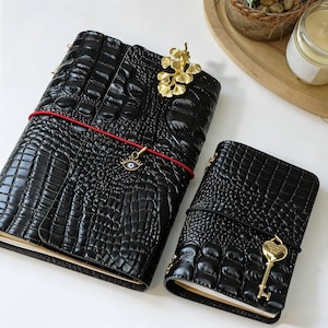May include: Two black leather journals with a crocodile skin texture. The larger journal has a red elastic band, a gold charm with an eye design, and a gold leaf-shaped clip. The smaller journal has a gold key charm.