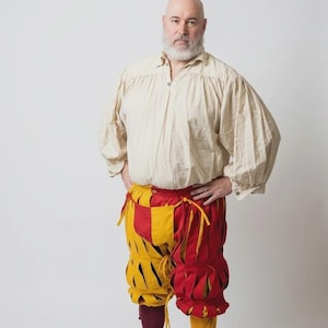 Peut inclure: Une chemise beige &agrave; manches longues avec un col boutonn&eacute; et des manches fronc&eacute;es, associ&eacute;e &agrave; une culotte &agrave; panneaux rouges et jaunes avec des fentes verticales et des liens. La tenue comprend une chaussette bordeaux et une jaune.