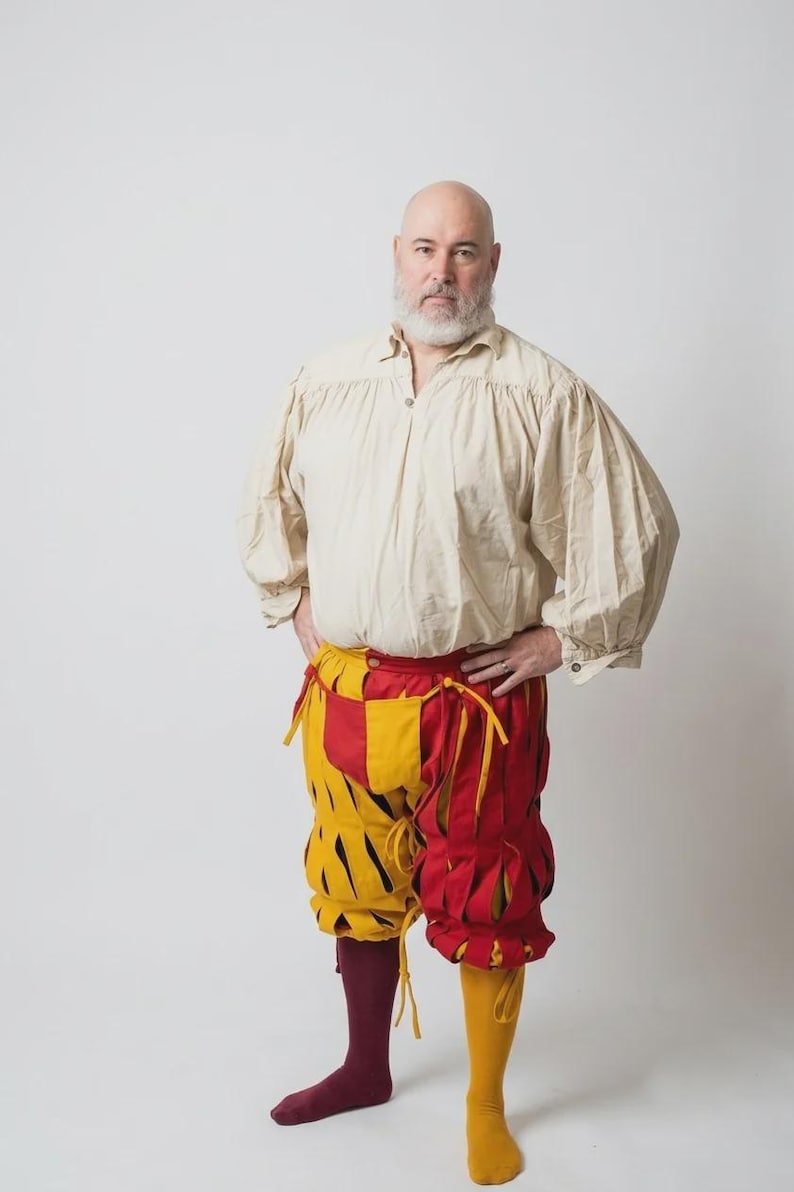 Peut inclure: Une chemise beige &agrave; manches longues avec un col boutonn&eacute; et des manches fronc&eacute;es, associ&eacute;e &agrave; une culotte &agrave; panneaux rouges et jaunes avec des fentes verticales et des liens. La tenue comprend une chaussette bordeaux et une jaune.