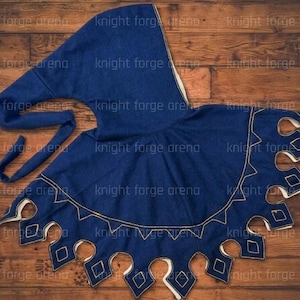 May include: A navy blue medieval hood and cape set. The hood has a long, pointed shape with a tie closure. The cape features a scalloped edge with diamond-shaped cutouts, and gold-colored stitching. The text "knight forge arena" is visible.