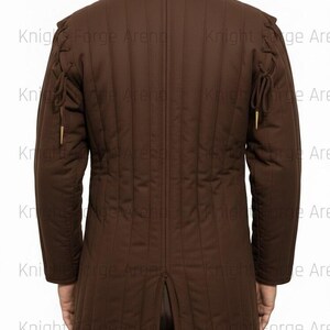 May include: Brown padded jacket with vertical stitching and long sleeves. Features decorative ties on the shoulders and sleeves. The jacket has a high collar and a split at the back. The text "Knight Forge Arena" is visible.