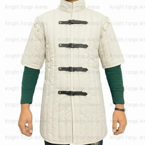 May include: Cream-colored, quilted medieval gambeson with short sleeves and a stand-up collar. The garment features four black leather straps with buckles down the front and is worn over a green long-sleeved shirt.