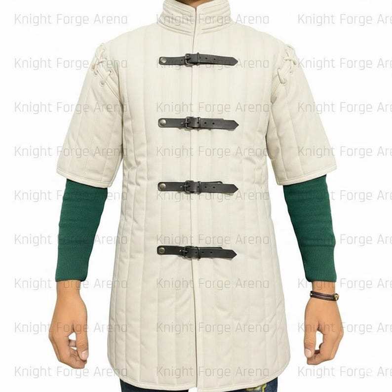 May include: Cream-colored, quilted medieval gambeson with short sleeves and a stand-up collar. The garment features four black leather straps with buckles down the front and is worn over a green long-sleeved shirt.