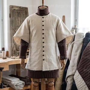 以下が含まれることがあります： 中世スタイルのチュニックと下着のセット。チュニックは、ボタン留めのフロントと短い袖が付いた、明るいベージュ色です。下着は濃い茶色です。この衣装は木製のマネキンに展示されています。