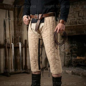 May include: Beige quilted medieval leg armor with black lacing details. The armor is worn with a brown leather belt and wrist cuff. A brown leather cord is held in the hand. The image is set in a medieval setting.
