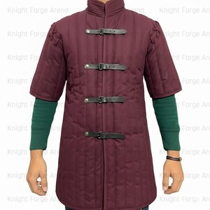 May include: A burgundy quilted jacket with short sleeves and a stand-up collar. The jacket features three black leather straps with buckles down the front. The sleeves are short, revealing green sleeves underneath. The jacket is part of a medieval or historical clothing collection.
