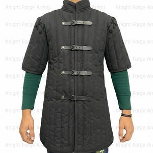 May include: Black padded gambeson with short sleeves and a stand-up collar. The medieval garment features three leather straps with buckles down the front. The sleeves are short, and the garment is quilted.