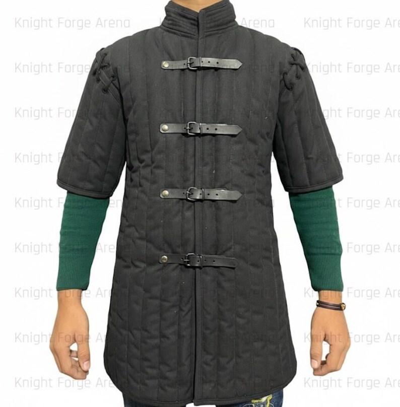 May include: Black padded gambeson with short sleeves and a stand-up collar. The medieval garment features three leather straps with buckles down the front. The sleeves are short, and the garment is quilted.