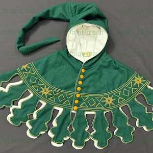May include: A green medieval hood and cape set. The hood has a long, pointed tip and a white interior. The cape features a scalloped edge with gold embroidery and yellow button closures. The text "Knight forge arena" is visible.