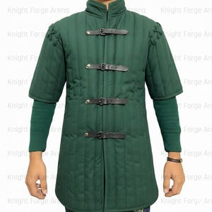 May include: A dark green padded gambeson with short sleeves and a stand-up collar. The front closure features three black leather straps with buckles. The sleeves are accented with decorative stitching. The gambeson is designed for historical reenactment or costume use.
