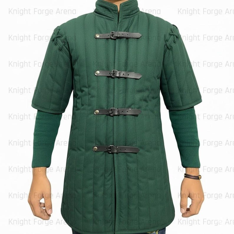 May include: A dark green padded gambeson with short sleeves and a stand-up collar. The front closure features three black leather straps with buckles. The sleeves are accented with decorative stitching. The gambeson is designed for historical reenactment or costume use.