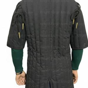 May include: Black padded gambeson with short sleeves and a high collar. The quilted fabric features vertical stitching. The sleeves have decorative gold-colored accents. The back of the garment is visible, showcasing the design and construction.